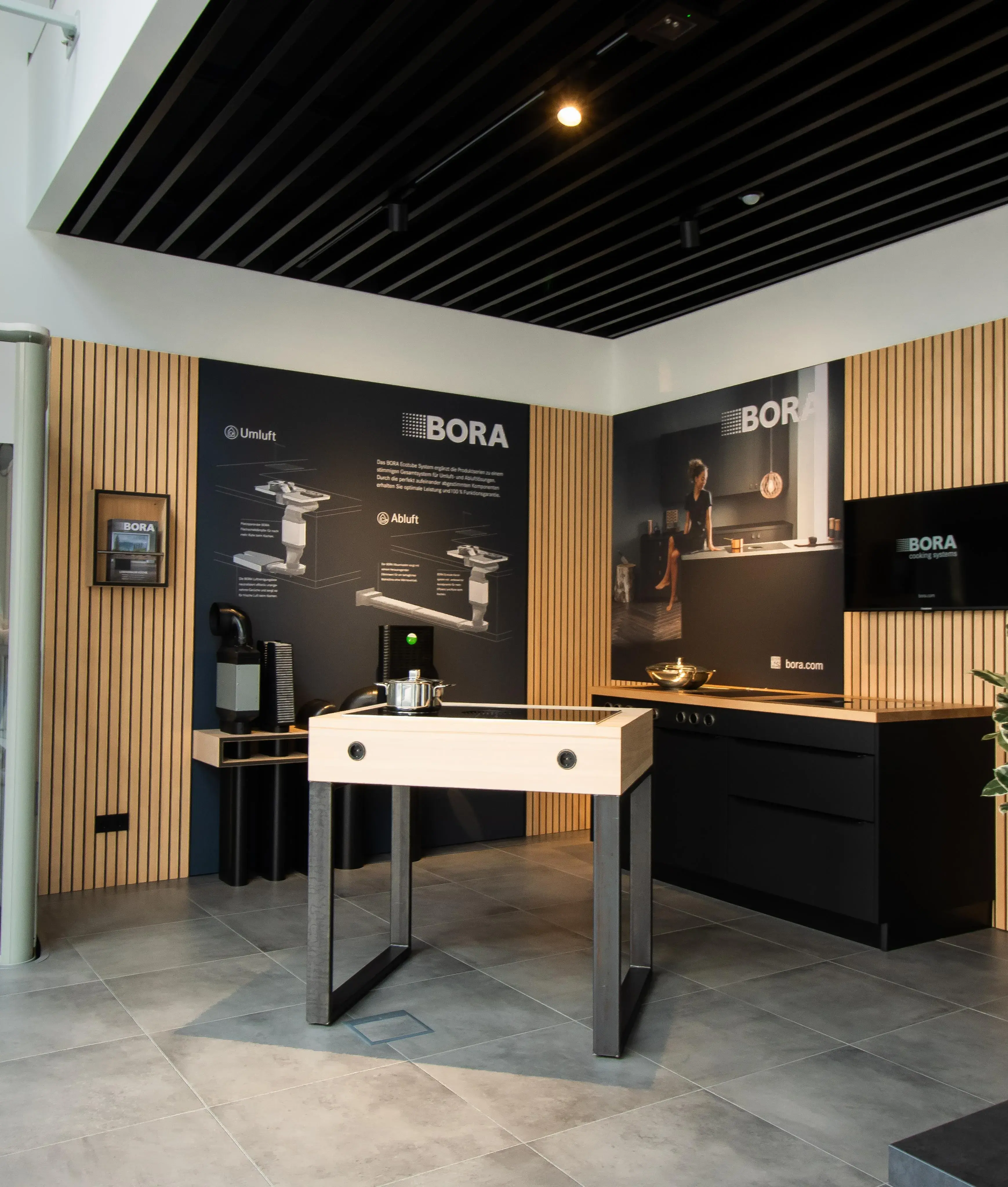 Moderne Küchenausstellung mit Holz- und Schwarzakzenten, zentraler Kochinsel und Infotafeln.